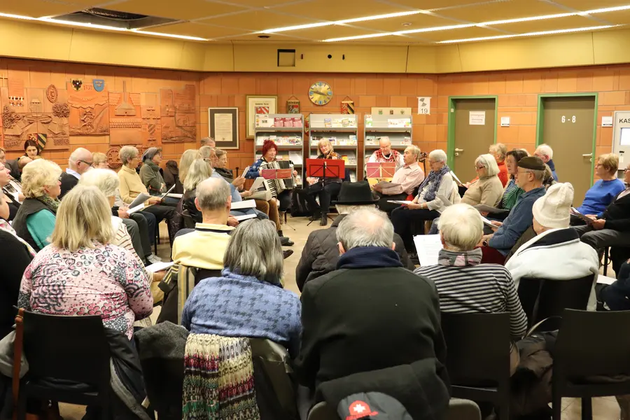 Sängerinnen und Sänger sitzen im Foyer des Rathauses im Kreis. In der Mitte Musikerinnen mit Geige, Akkordeon und Gitarre. 