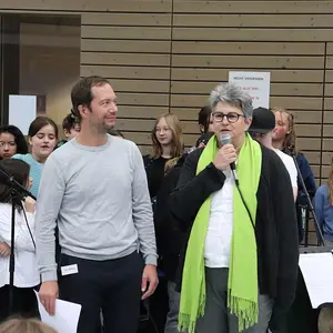 Eine Frau spricht in ein Mikrofon, daneben ein Mann und im Hintergrund einige Schüler