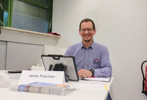 Jens Fischer sitzt vor seinem aufgestellten Laptop. Vor ihm Broschüren und sein Namensschild. 
