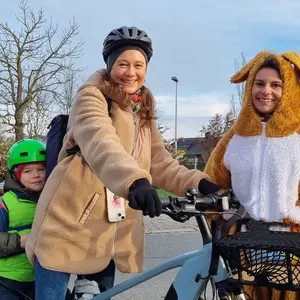 eine Mutter mit einem Lastenrad, hinter ihr sitzen ihre beiden Kinder, neben ihr steht eine Frau im Hasenkostüm 