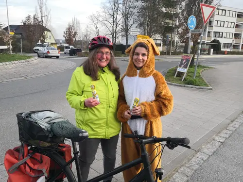 Eine Frau mit Fahrrad samt Packtasche, Warnweste und Fahrradhelm. Neben ihr eine Frau im Plüschhasenkostüm. Beide halten Schokohasen in den Händen. 