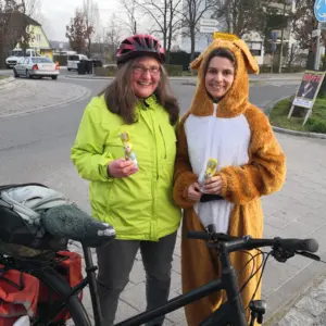 Eine Frau mit Fahrrad samt Packtasche, Warnweste und Fahrradhelm. Neben ihr eine Frau im Plüschhasenkostüm. Beide halten Schokohasen in den Händen. 