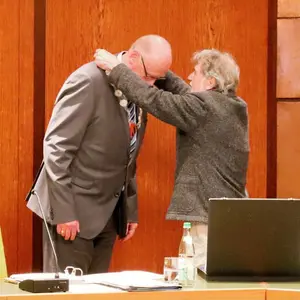 Zwei Männer vor Holzwand. Der rechts stehende legt dem Linken die Amtskette aus Silber um. 