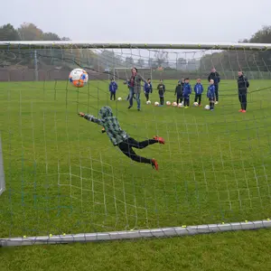 Menschen auf einem Rasen-Fußballfeld. Man schaut von hinten durch ein Tornetz. Der Torwart versucht gerade, einen Ball zu fangen. 