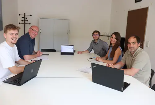 Vier Männer und eine Frau sitzen mit Laptops in einem Besprechungszimmer und schauen in die Kamera.