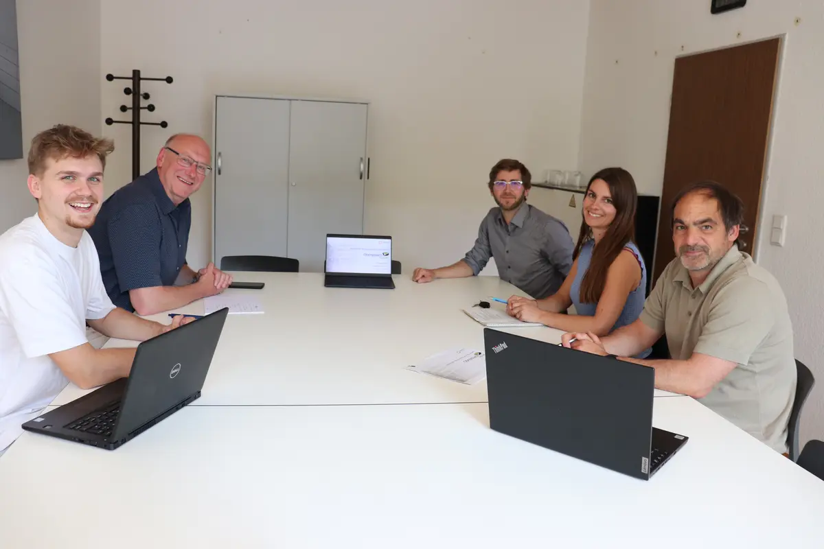 Vier Männer und eine Frau sitzen mit Laptops in einem Besprechungszimmer und schauen in die Kamera.