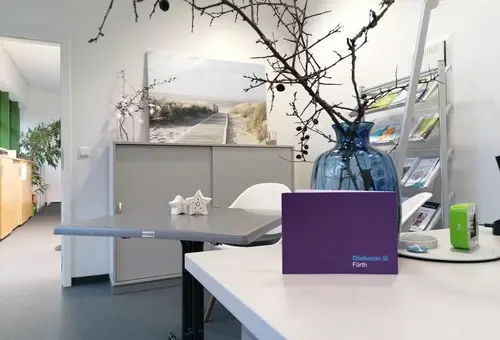 Im Vordergrund Teile des Schreibtisches mit Blume und im Hintergrund ein Besprechungstisch mit Büromöbeln.