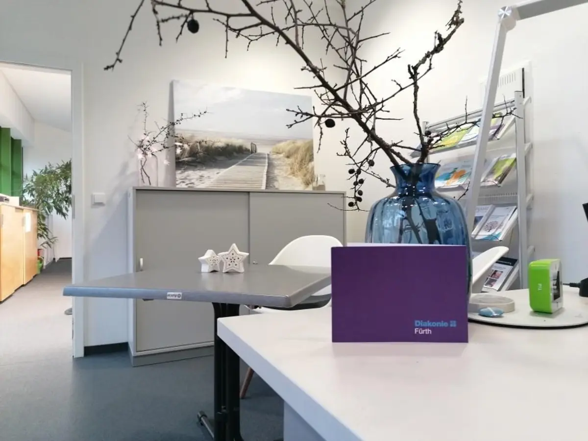 Im Vordergrund Teile des Schreibtisches mit Blume und im Hintergrund ein Besprechungstisch mit Büromöbeln.