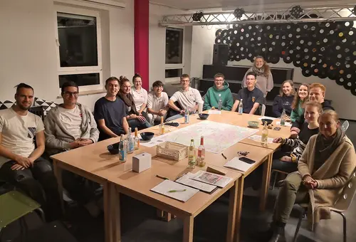 Um einen Tisch sitzen rund 16 Personen herum