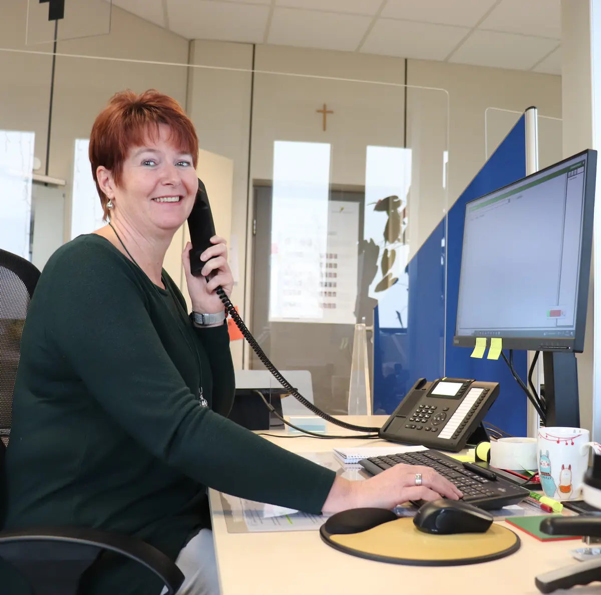 Sandra Bernard an ihrem Arbeitsplatz vor dem Bildschirm, in der Hand einen Telefonhörer 