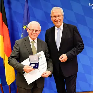 Zwei Männer mit der Medaille vor den Flaggen Bayern, Deutschlands und Europas im Hintergrund.