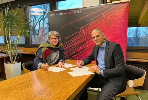 Eine Frau und ein Mann sitzen an einem Tisch und unterschreiben Papiere vor einem Banner der Deutschen Telekom