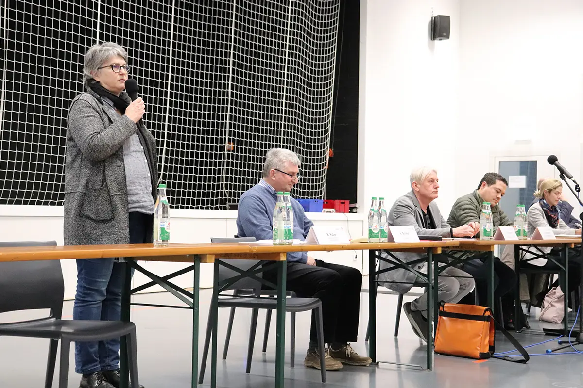 Eine Frau spricht stehend ins Mikrofon und fünf weitere Personen sitzend am Podium.