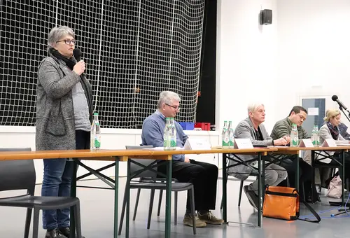 Eine Frau spricht stehend ins Mikrofon und fünf weitere Personen sitzend am Podium.