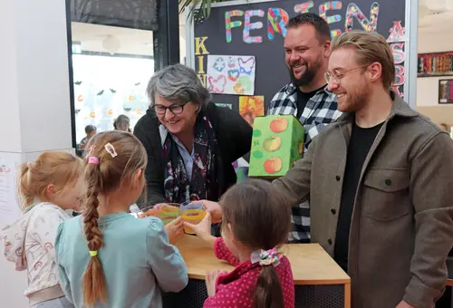 Birgit Huber und Mitarbeiter des Horts mit einer Kiste Apfelsaft. Im Vordergrund 3 Mädchen, die mit Saftgläsern anstoßen. 