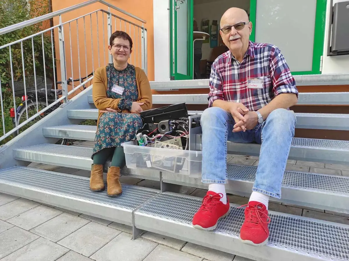 Eine Frau und ein älterer Herr sitzen auf den Stufen vor dem Oberasbacher Jugendhaus, zwischen sich ein kaputter Kaffeevollautomat.