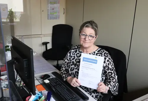 Ute Schmidt-Alpers am Schreibtisch mit Zertifikat der Rentenversicherung in der Hand. 