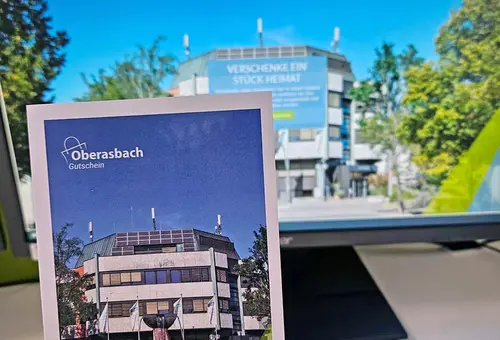 Eine Frauenhand hält eine Geschenkkarte mit dem neuen Oberasbach-Gutschein, im Hintergrund ist die dazugehörige Webseite auf einem Bildschirm zu sehen