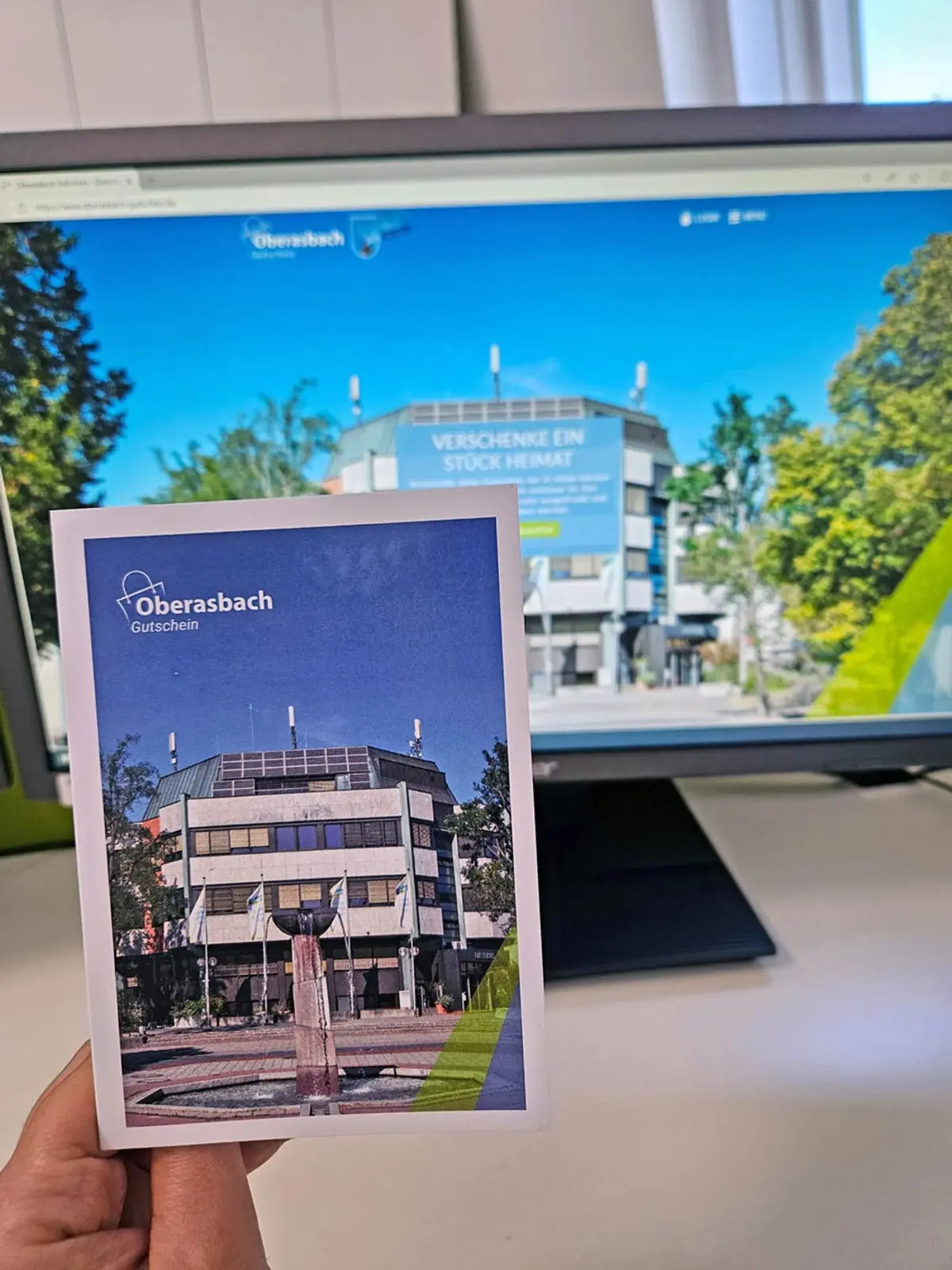 Eine Frauenhand hält eine Geschenkkarte mit dem neuen Oberasbach-Gutschein, im Hintergrund ist die dazugehörige Webseite auf einem Bildschirm zu sehen