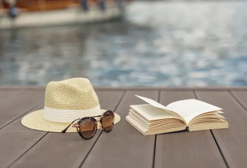 Strohhut, Sonnenbrille und offenes Buch am Rand eines Swimmingpools 