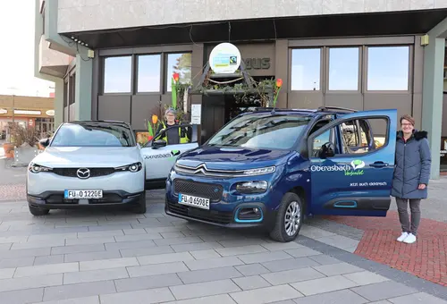 Zwei Frauen stehen neben einem vor dem Rathaus geparkten weißen und einem dunkelblauen Auto, die jeweils Aufkleber mit dem Logo "Oberasbach verbindet" und dem Zusatz "jetzt auch elektrisch" auf den Türen tragen. 