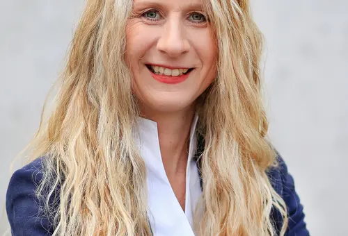 Frau mit langen blonden Locken, weißer Bluse und blauer Anzugjacke 