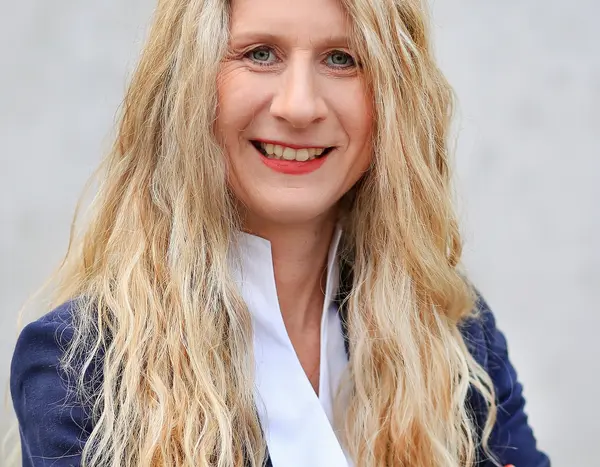 Frau mit langen blonden Locken, weißer Bluse und blauer Anzugjacke 