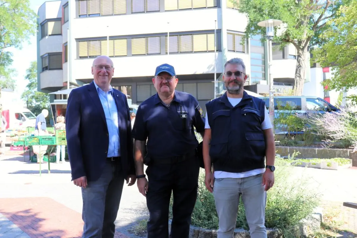 Drei Männer vor dem Oberasbacher Rathaus, im Hintergrund ist ein Wochenmarkt zu sehen