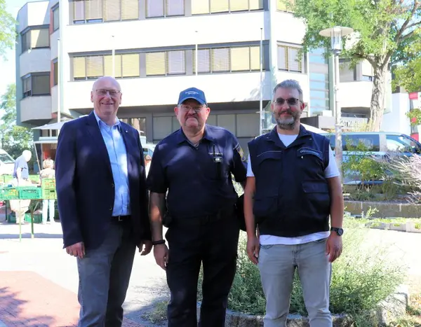 Drei Männer vor dem Oberasbacher Rathaus, im Hintergrund ist ein Wochenmarkt zu sehen