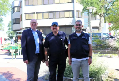 Drei Männer vor dem Oberasbacher Rathaus, im Hintergrund ist ein Wochenmarkt zu sehen