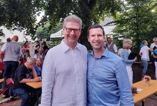 Zwei Männer schauen in die Kamera. Im Hintergrund Bierbänke mit weiteren Gästen.