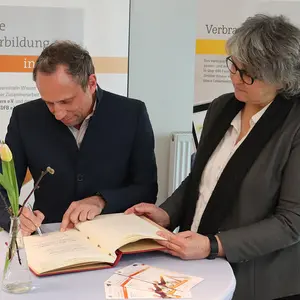 Ein Mann schriebt in das Buch, eine Frau schaut ihm zu.