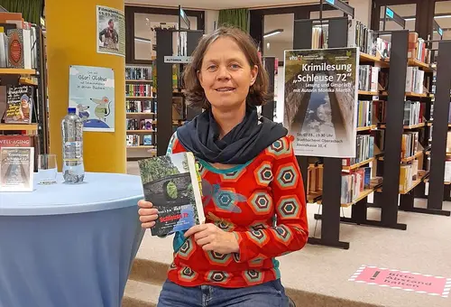 Die Autorin Monika Martin hält ein Buch in die Kamera, im Hintergrund sind Bücherregale und das Plakat zur Lesung zu sehen.