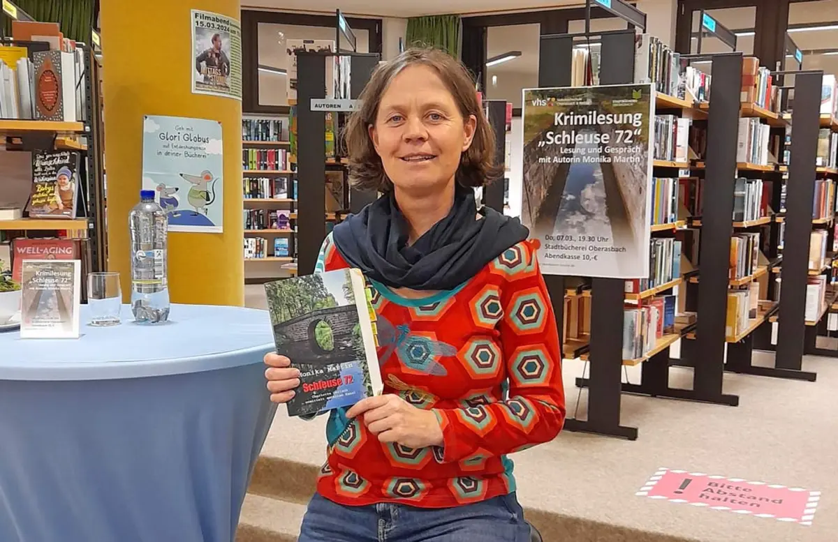 Die Autorin Monika Martin hält ein Buch in die Kamera, im Hintergrund sind Bücherregale und das Plakat zur Lesung zu sehen.