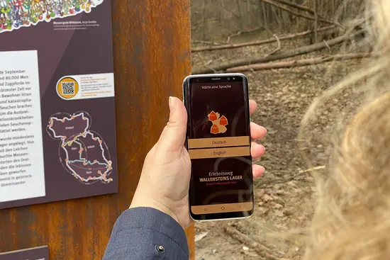 Eine Frauenhand hält ein Handy an den QR-Code der Stele. Das Display zeigt die App Erlebnisweg Wallensteins Lager an