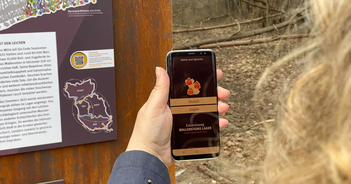Eine Frauenhand hält ein Handy an den QR-Code der Stele. Das Display zeigt die App Erlebnisweg Wallensteins Lager an
