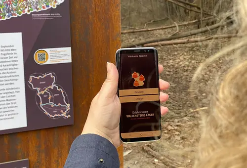 Eine Frauenhand hält ein Handy an den QR-Code der Stele. Das Display zeigt die App Erlebnisweg Wallensteins Lager an