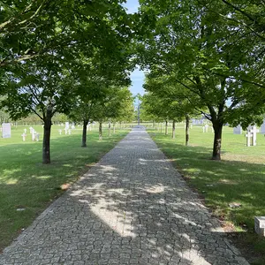 Ein Weg führt über eine grüne Wiese, gesäumt von Bäumen. Auf der Wiese stehen weiße Grabsteine.