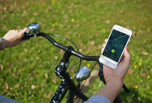 Blick von oben auf einen Fahrradlenker. Das Fahrrad steht auf einer grünen Wiese. Der/die Lenkende hält ein Handy mit der Stadtradeln-App in der Hand. hält in der rechten 
