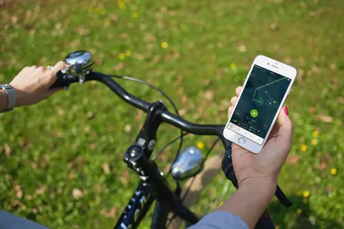 Blick von oben auf einen Fahrradlenker. Das Fahrrad steht auf einer grünen Wiese. Der/die Lenkende hält ein Handy mit der Stadtradeln-App in der Hand. hält in der rechten 