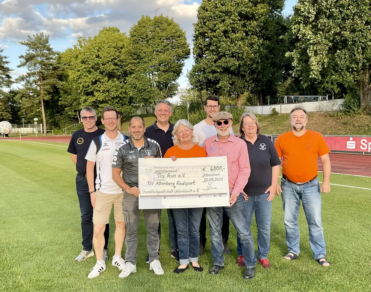 9 Personen beim Gruppenfoto auf einem Sportplatz mit einem großen Scheck über 6.000 EUR in der Hand.