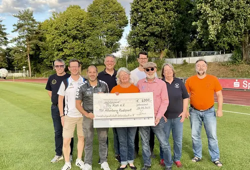 9 Personen beim Gruppenfoto auf einem Sportplatz mit einem großen Scheck über 6.000 EUR in der Hand.
