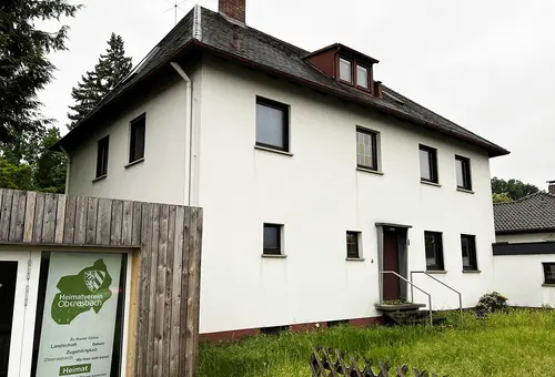 Haus mit hohem Gras im Vorgarten. Links Teile der Garage mit dem Logo des Heimatvereins.
