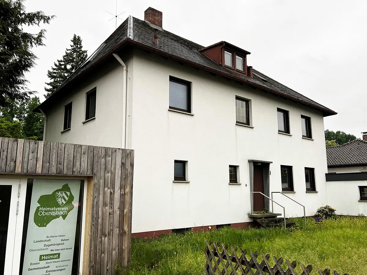 Haus mit hohem Gras im Vorgarten. Links Teile der Garage mit dem Logo des Heimatvereins.