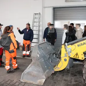 Bauhof-Mitarbeiter in oranger Sicherheitskleidung und eine Gruppe Jugendlicher. Im Vordergrund die Schaufel eines Baggers. 