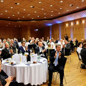 Blick in einen Raum mit gedeckten runden Tischen und festlich gekleideten Gästen.