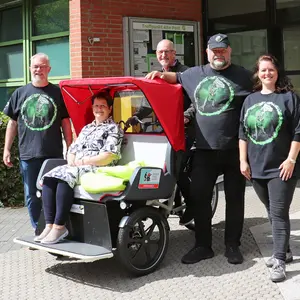 Eine Frau sitzt in einer E-Rikscha. Darum herum stehen drei Männer und eine Frau mit T-Shirts der United Kiltrunners.  