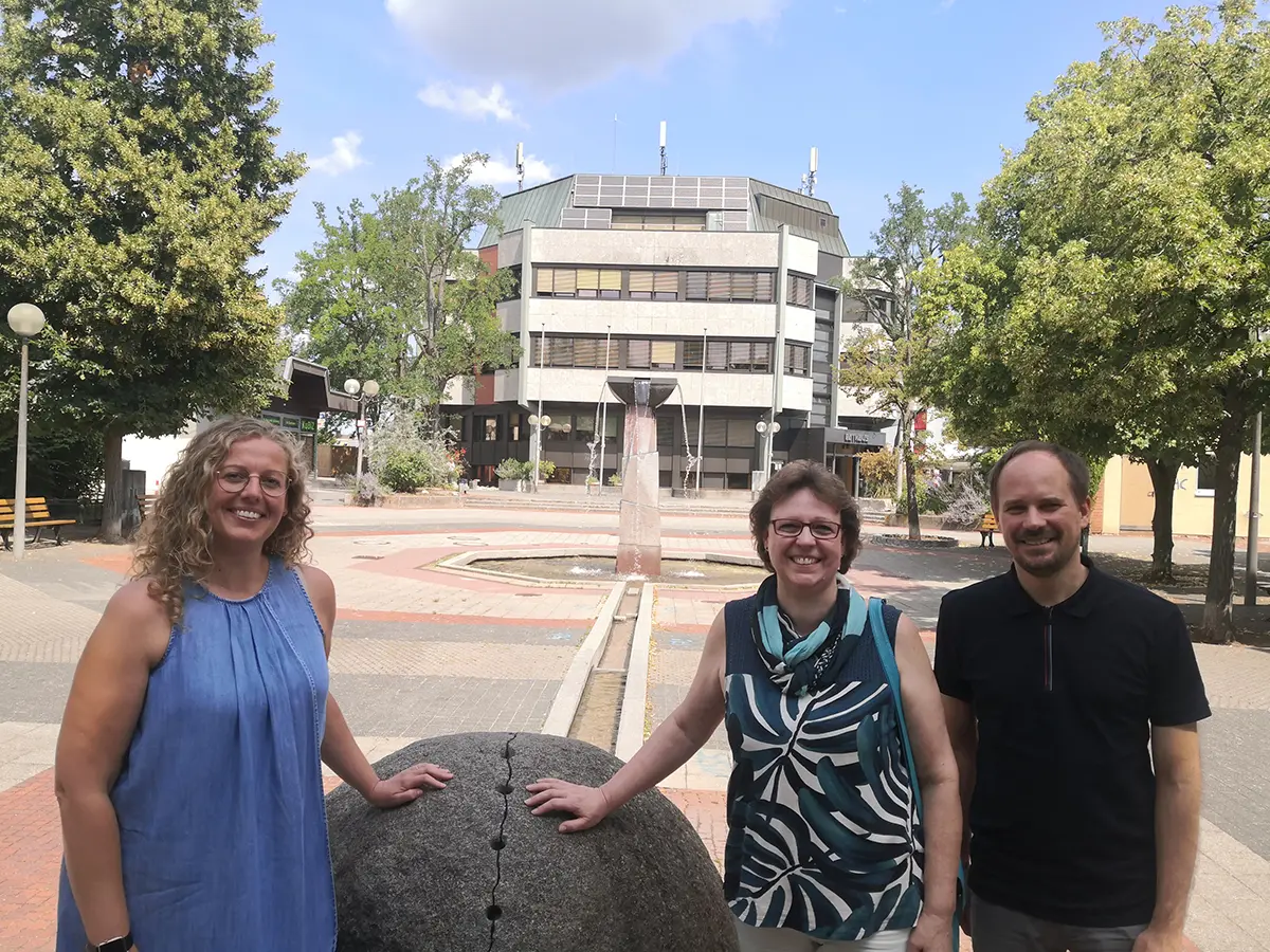 Zwei Frauen und ein Mann an der großen Steinkugel am Brunnen auf dem Rathausplatz. Im Hintergrund das Rathaus.
