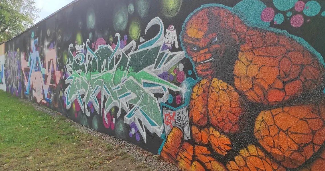 Ein hulkartiger, orange-brauner, glatzköpfiger Muskelmann neben einem Tag