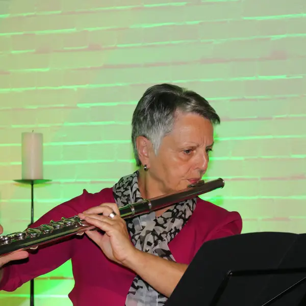 Frau mit Querflöte und Notenständer. Im Hintergrund eine grüne Wand und ein Kerzenleuchter mit zwei Kerzen. 